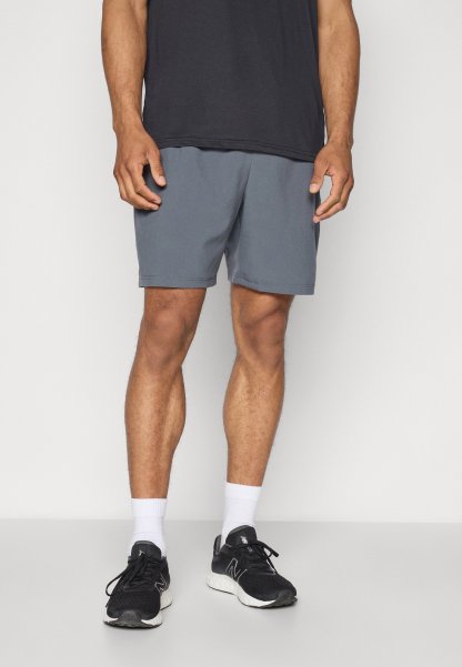 Hombre vistiendo pantalones cortos grises, zapatillas negras, calcetines blancos y una camiseta negra, de pie contra un fondo blanco sencillo.