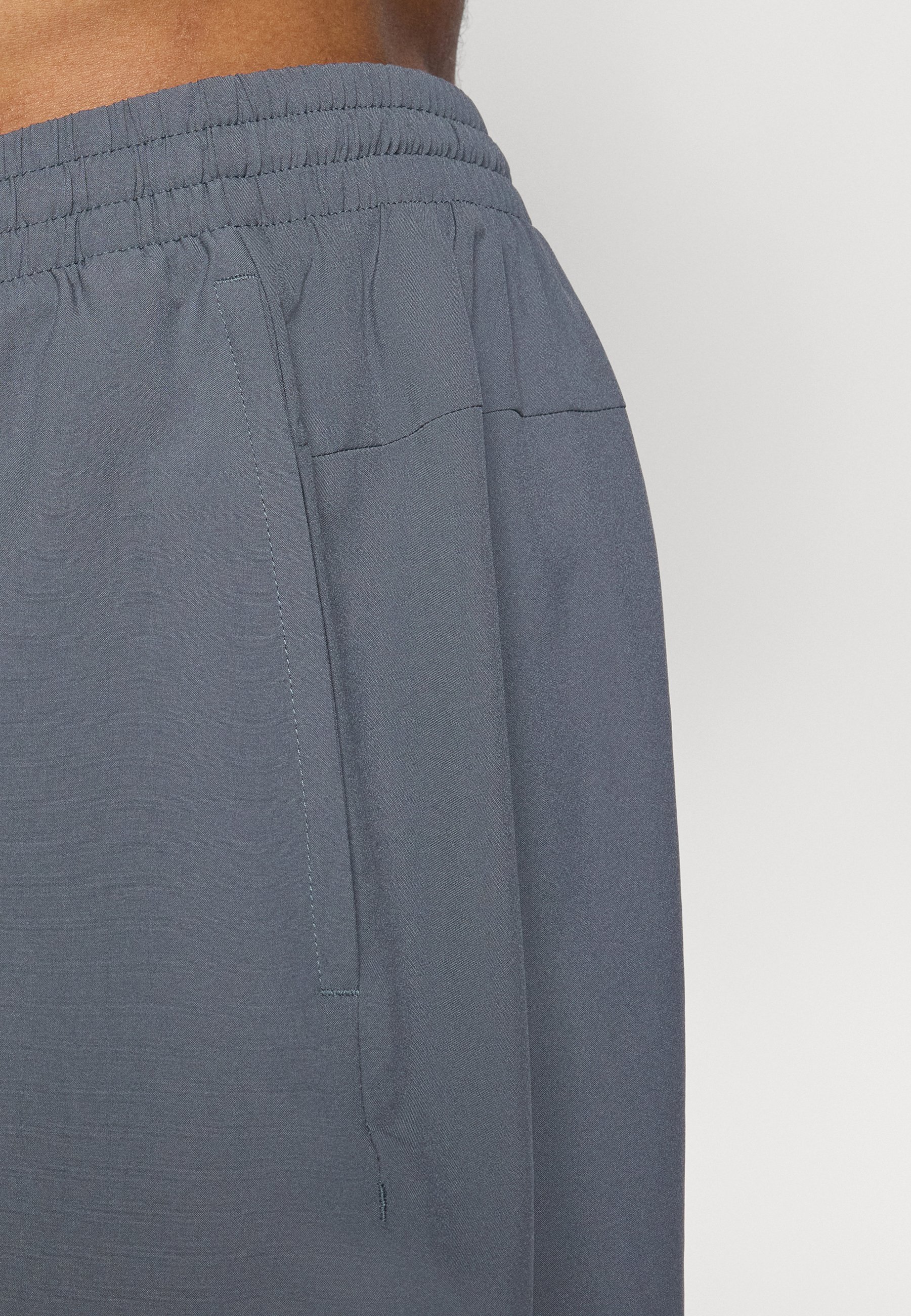 Acercamiento a la cintura de una persona que lleva pantalones con cinturilla elástica de color gris oscuro y un bolsillo lateral, contra un fondo claro y liso.