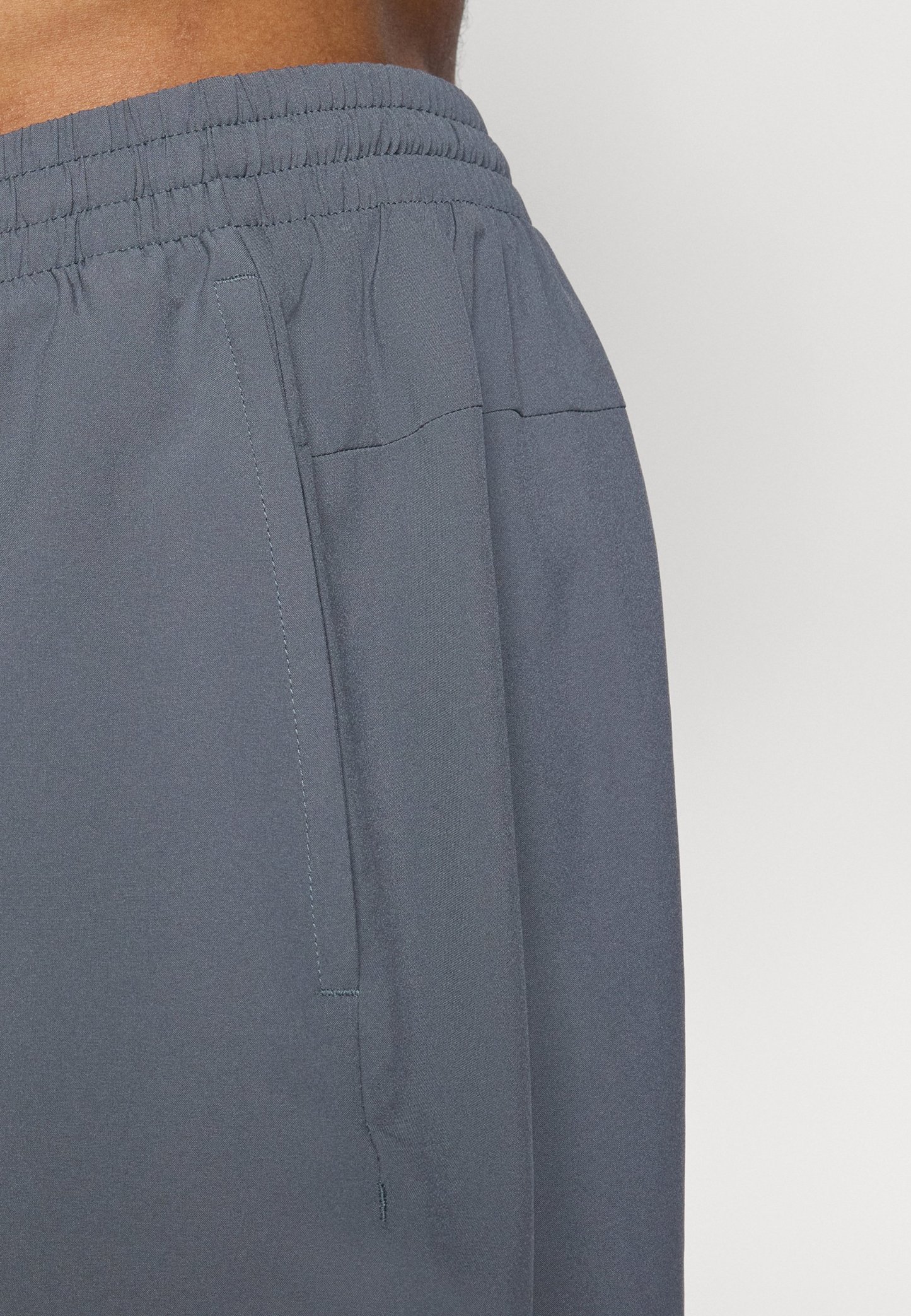 Acercamiento a la cintura de una persona que lleva pantalones con cinturilla elástica de color gris oscuro y un bolsillo lateral, contra un fondo claro y liso.
