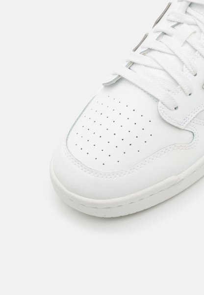 Zapatilla blanca de cuero con caja de dedos perforada, cordones blancos planos y suela de goma de color claro. Diseño redondeado con detalles de costura mínimos.