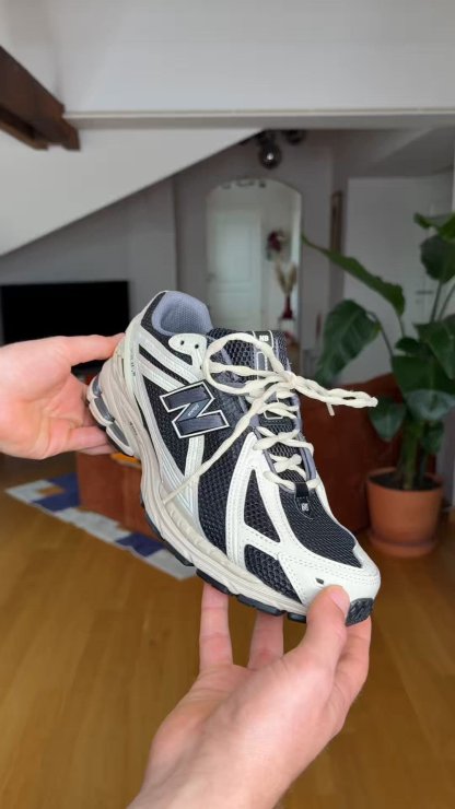 Manos sosteniendo una zapatilla para correr New Balance en blanco y negro con cordones blancos en una sala de estar con plantas y muebles.