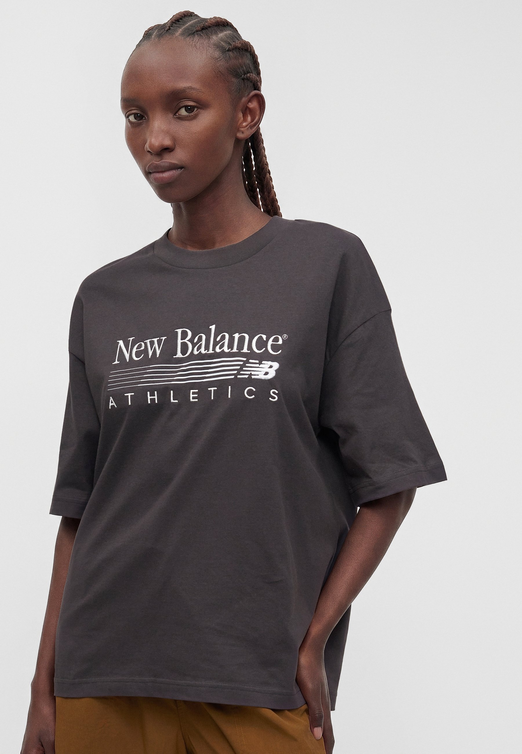 Mujer con cabello trenzado que lleva una camiseta holgada gris oscuro de New Balance Athletics y pantalones marrones, posando con una mano en el bolsillo.