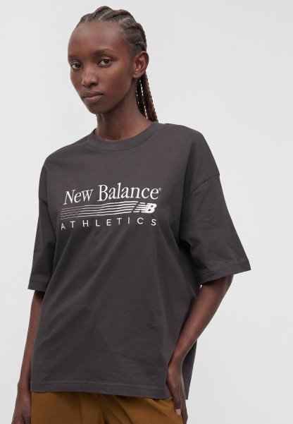 Mujer con cabello trenzado que lleva una camiseta holgada gris oscuro de New Balance Athletics y pantalones marrones, posando con una mano en el bolsillo.