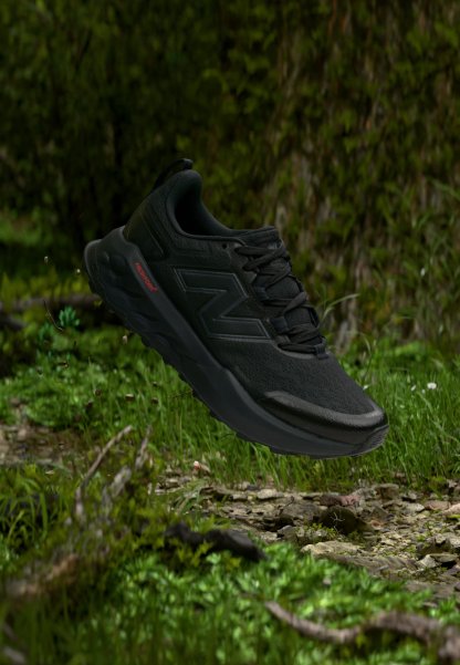 Zapatilla deportiva negra con suela gruesa y logo "N", flotando sobre un suelo forestal rocoso cubierto de musgo con vegetación al fondo.