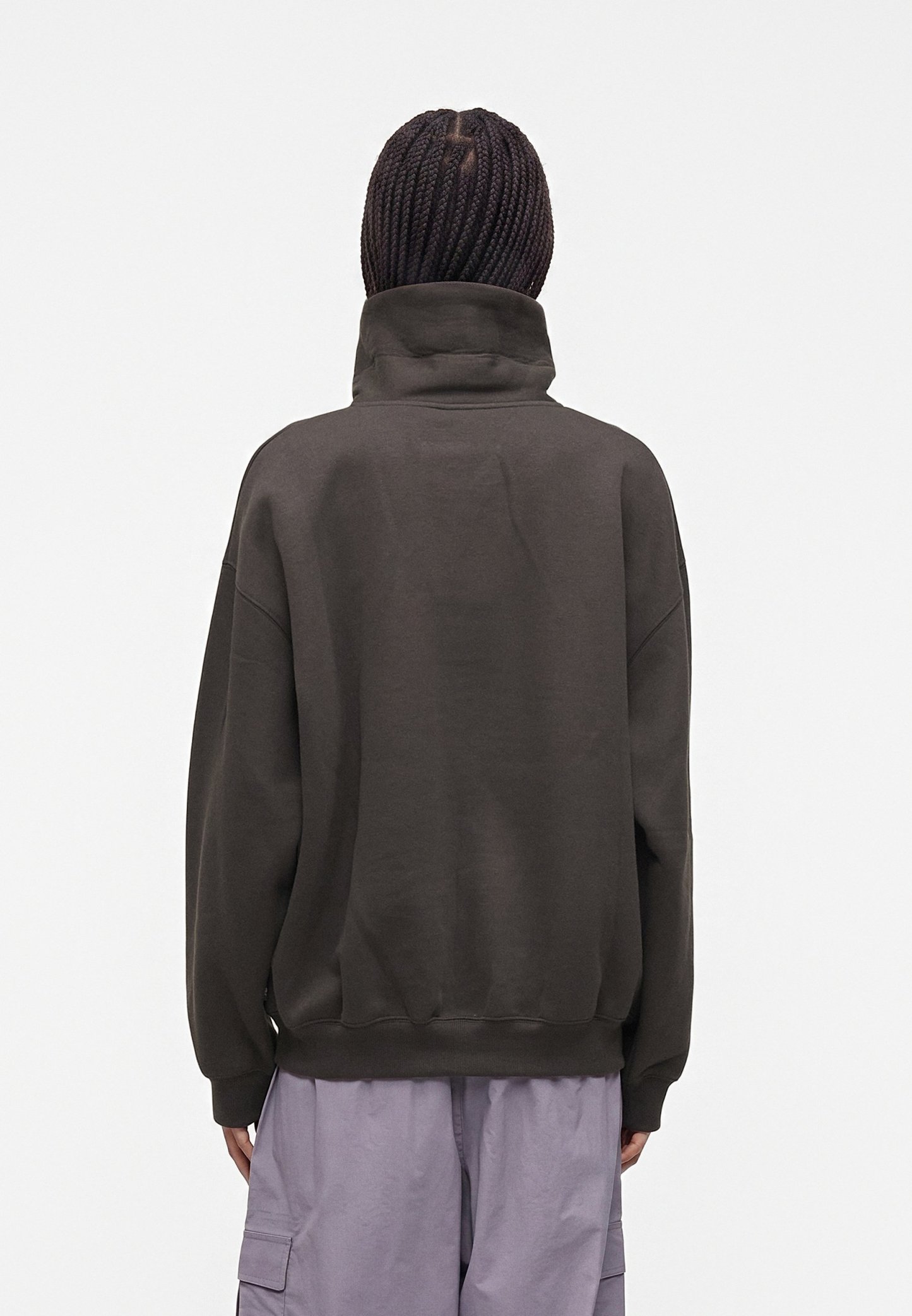 Persona con el pelo trenzado vista desde atrás, que lleva una sudadera oscura de cuello alto y pantalones cargo color lila claro sobre un fondo liso.