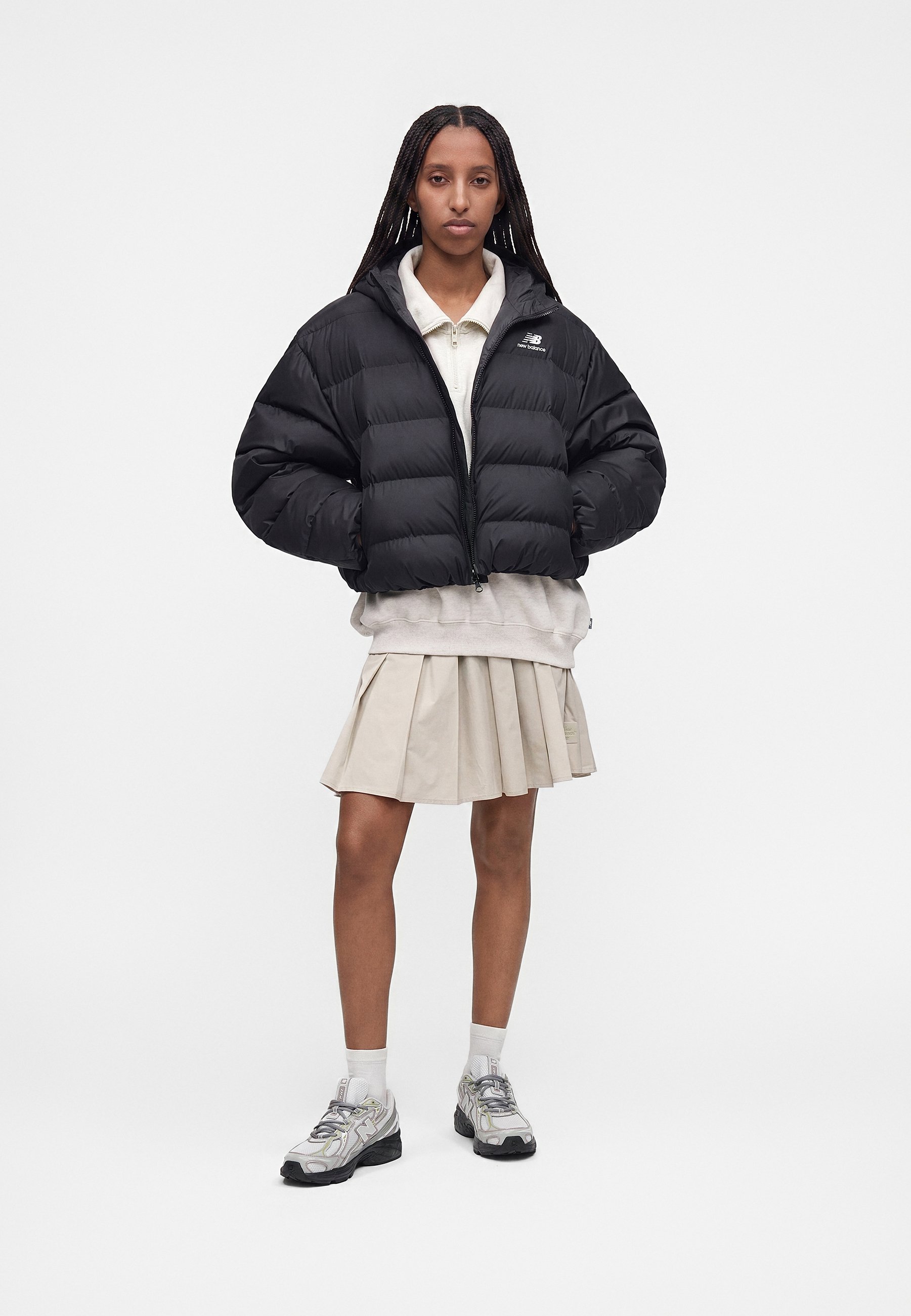 Mujer de pie con chaqueta acolchada negra, falda plisada beige, camiseta polo blanca, calcetines blancos y zapatillas grises sobre un fondo claro y liso.