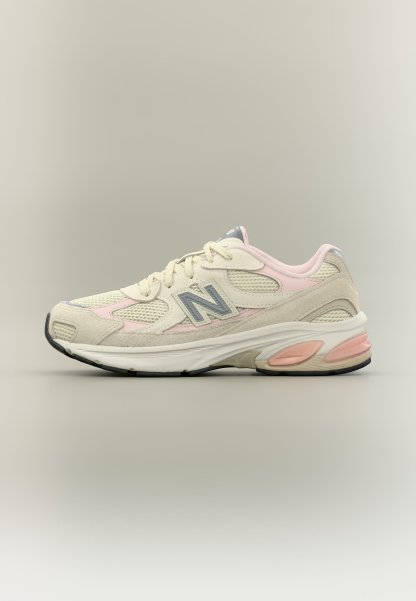 Zapatilla New Balance de color beige y rosa pálido con paneles de malla, superposiciones de ante y suela acolchada vista desde el lado izquierdo sobre un fondo neutro.