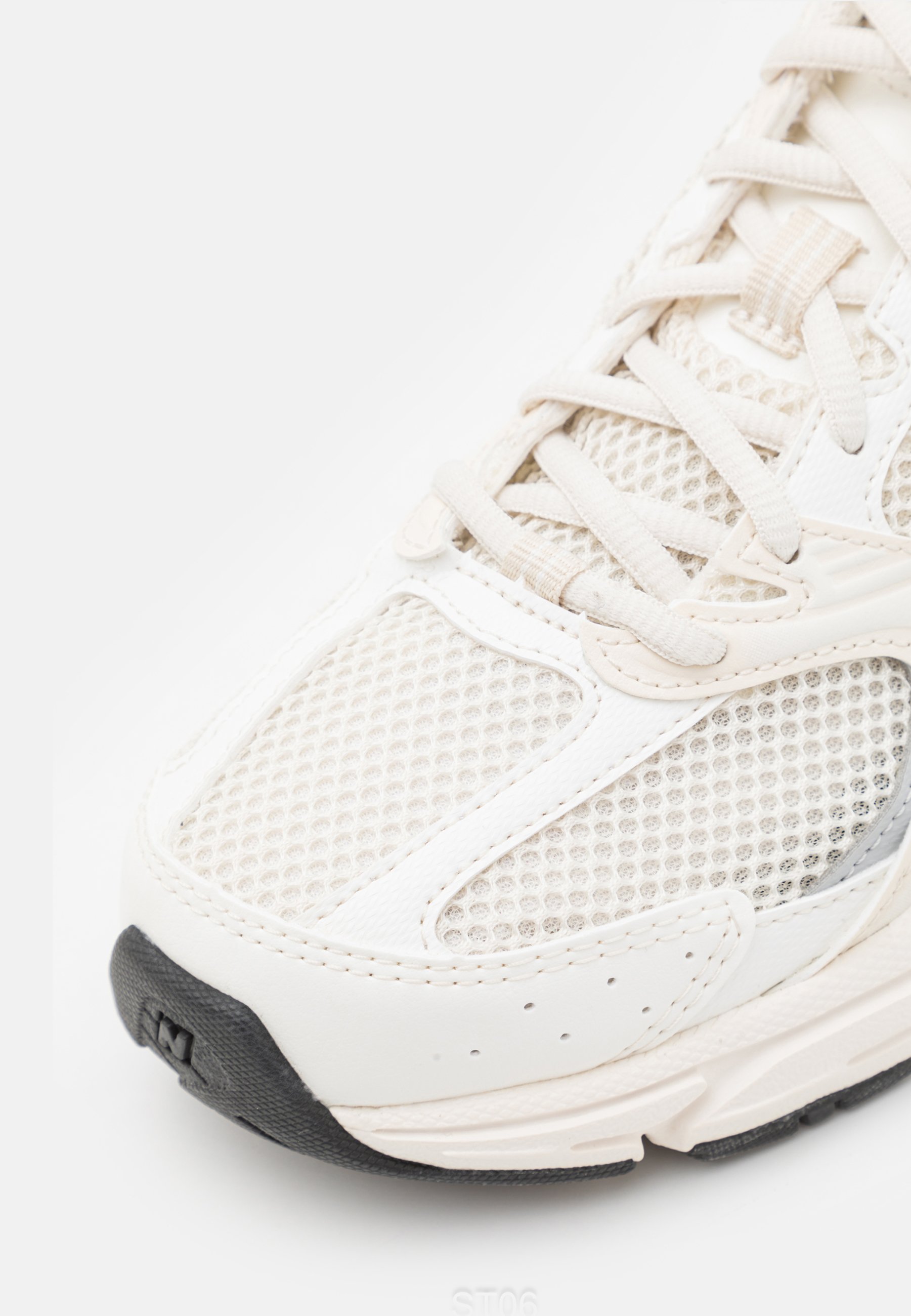Primer plano de un zapato deportivo blanco con paneles de malla, superposiciones de cuero, cordones blancos y una suela de goma negra en la punta.