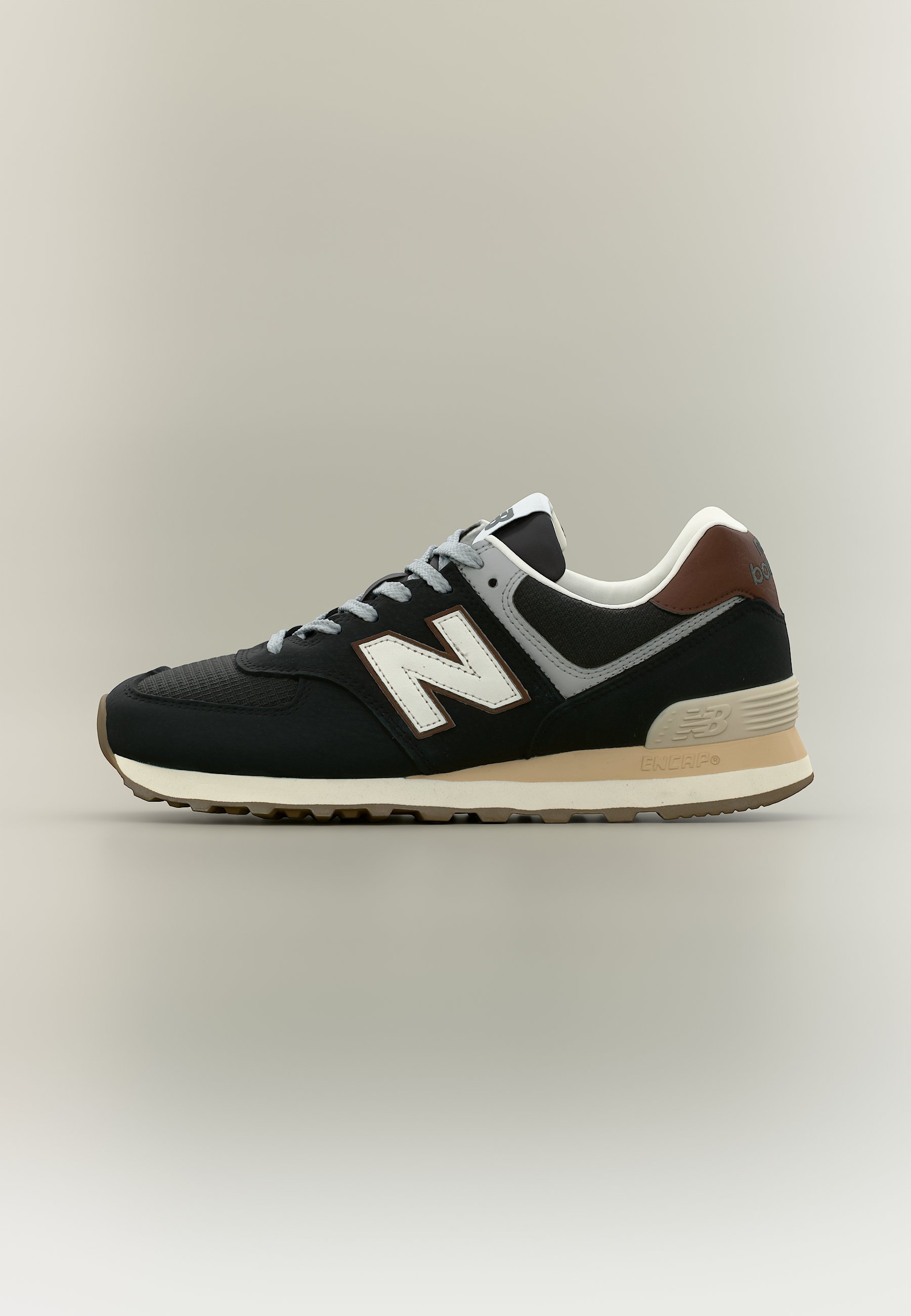 Zapatilla New Balance negra y gris con detalles en blanco y marrón, suela beige y cordones grises, vista lateral sobre un fondo neutro.
