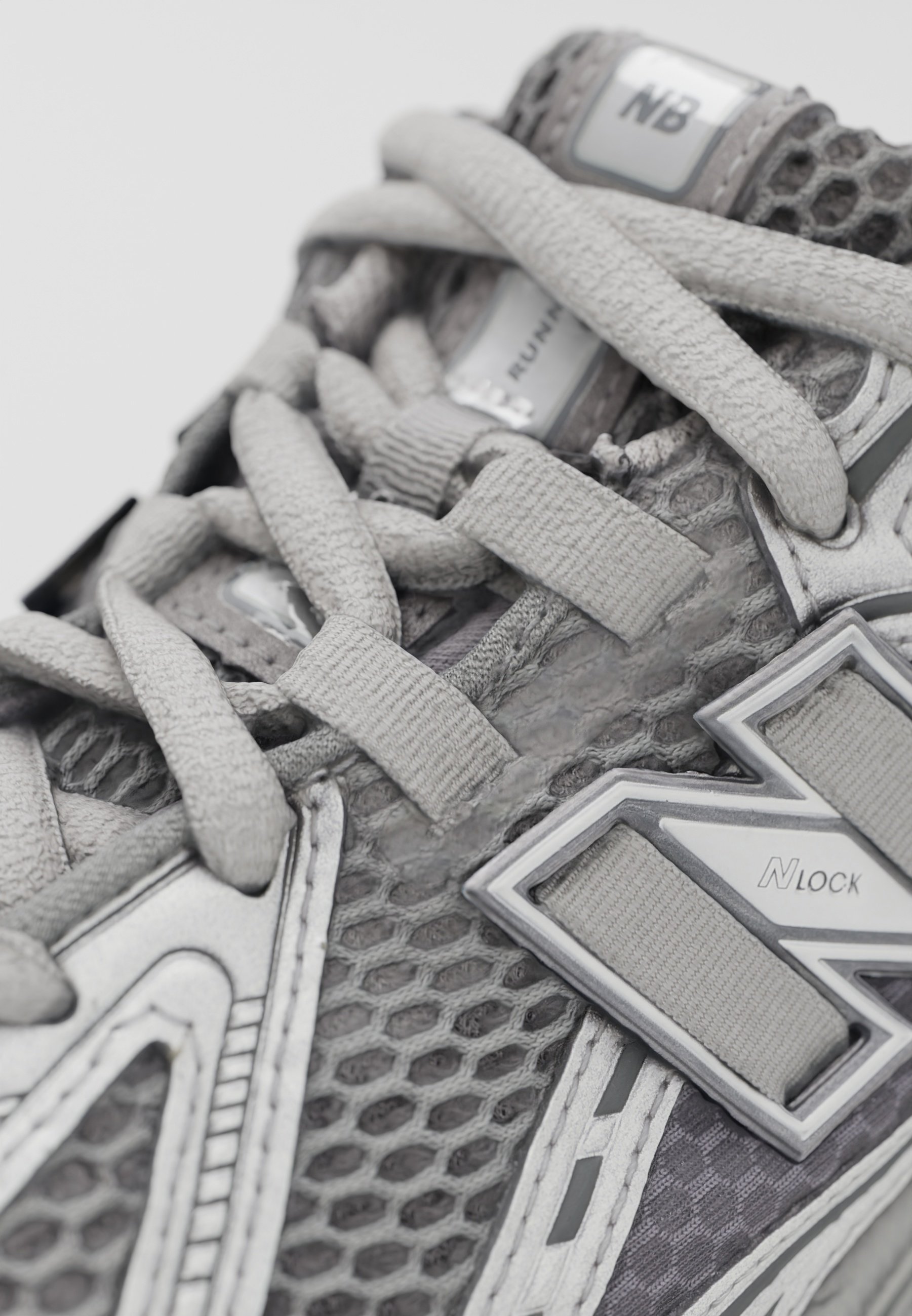Zapato deportivo gris con parte superior de malla, cordones gruesos y acentos metálicos. Los detalles notables incluyen patrones hexagonales y elementos reflectantes.