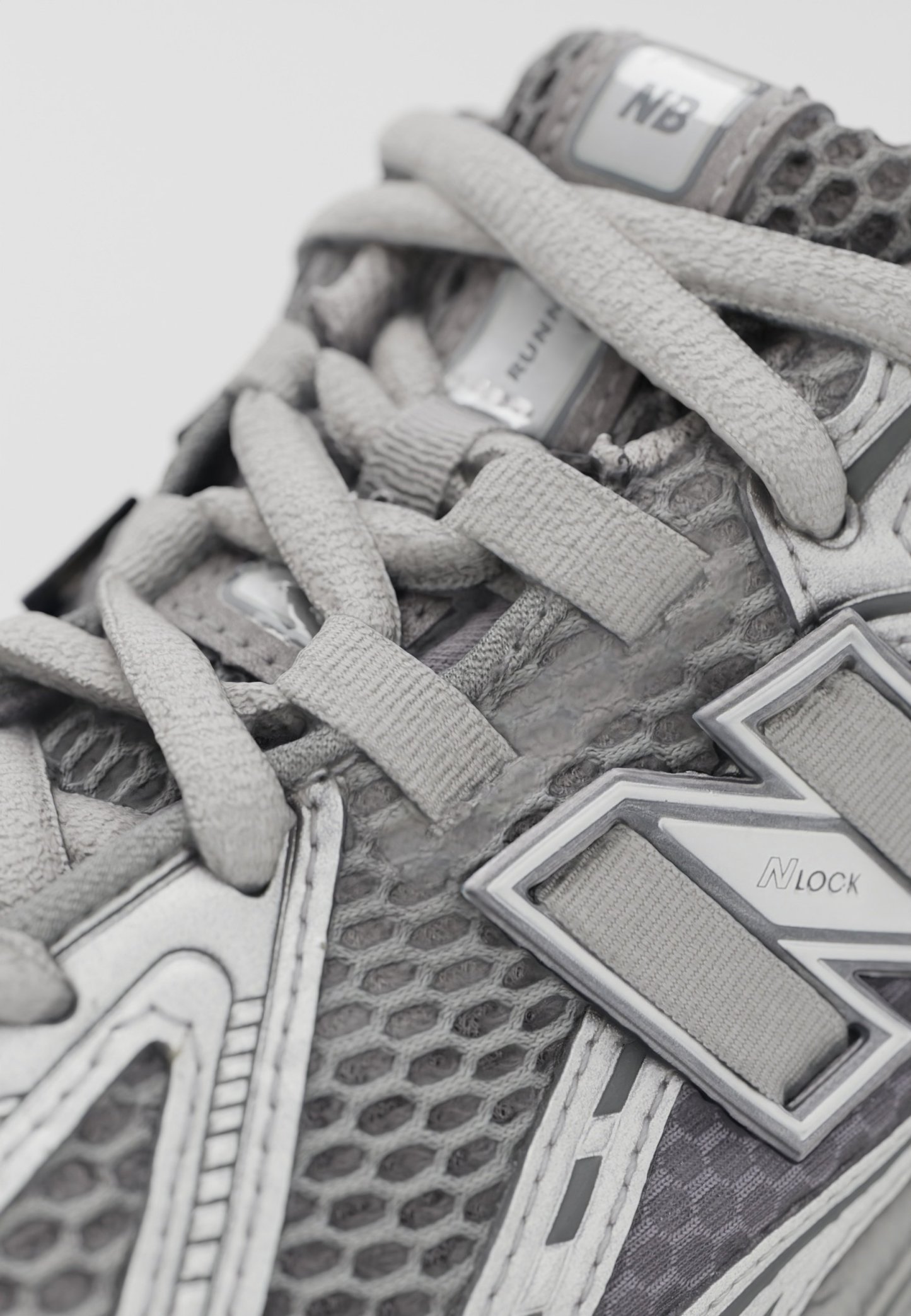 Zapato deportivo gris con parte superior de malla, cordones gruesos y acentos metálicos. Los detalles notables incluyen patrones hexagonales y elementos reflectantes.