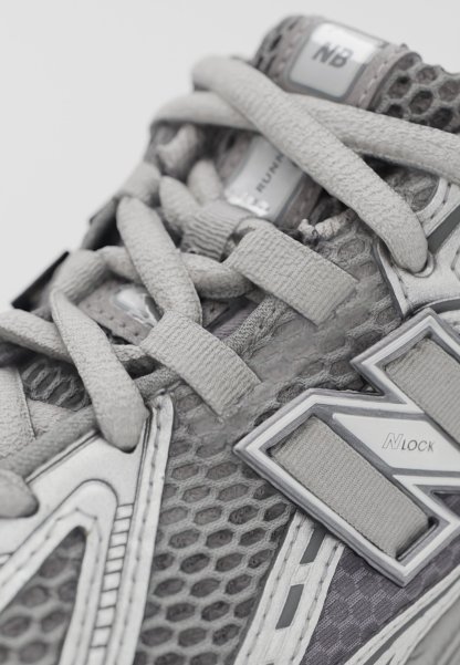 Zapato deportivo gris con parte superior de malla, cordones gruesos y acentos metálicos. Los detalles notables incluyen patrones hexagonales y elementos reflectantes.