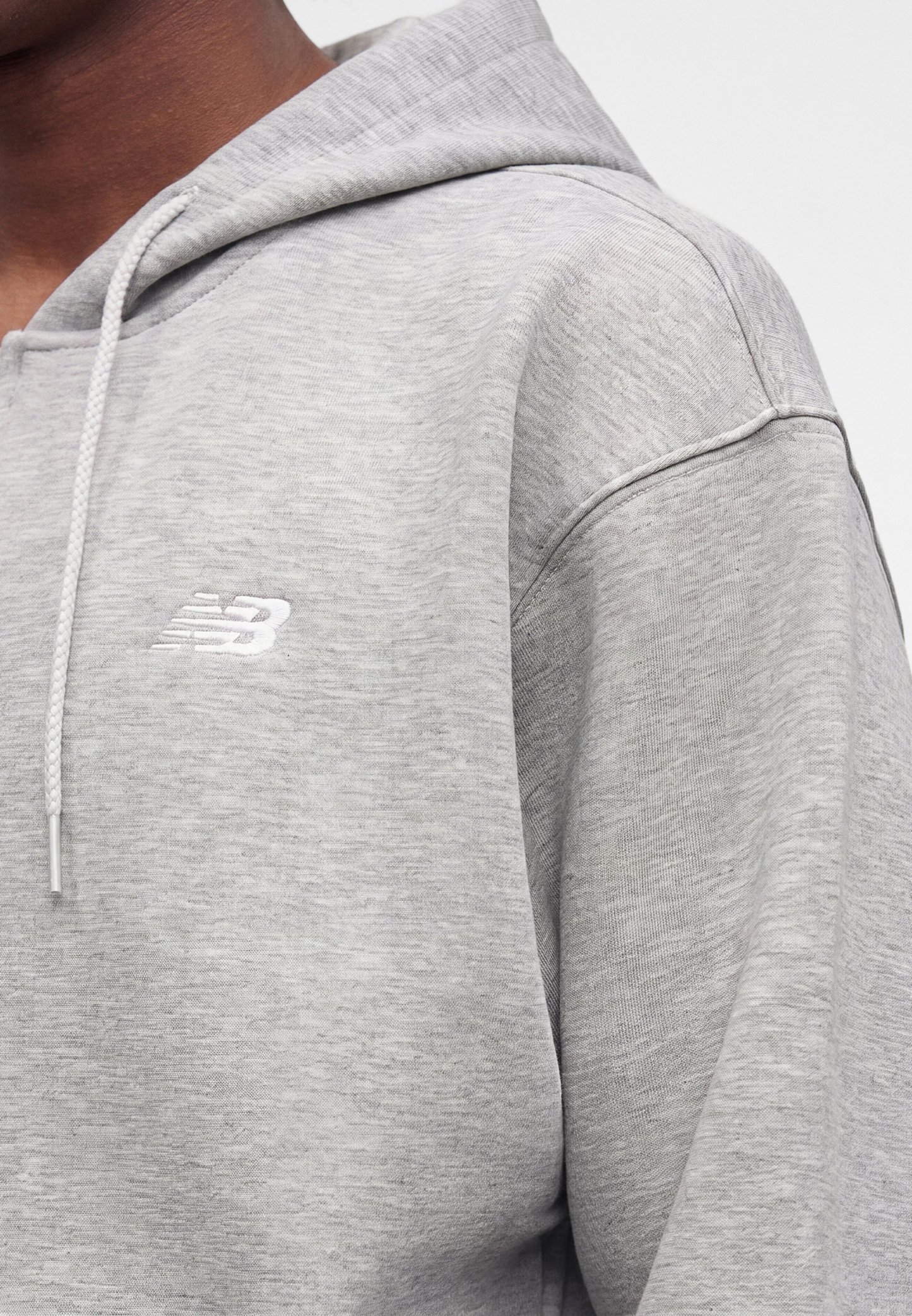 Persona con sudadera con capucha New Balance gris claro con cordón, mostrando la parte superior del pecho, el hombro y parte del cuello sobre un fondo blanco.
