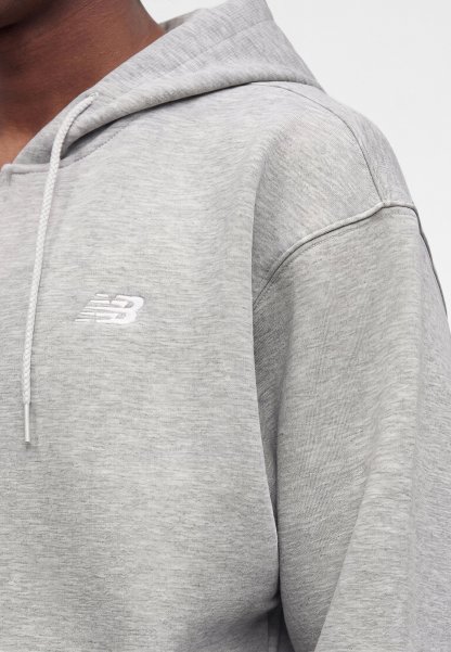Persona con sudadera con capucha New Balance gris claro con cordón, mostrando la parte superior del pecho, el hombro y parte del cuello sobre un fondo blanco.