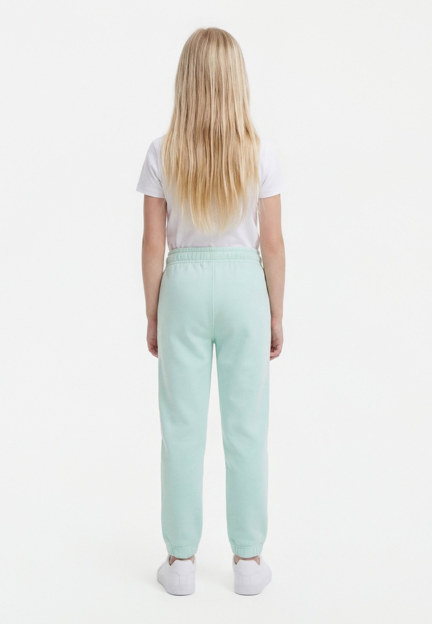 Niño con cabello largo y rubio, vistiendo una camiseta blanca, pantalones de chándal azul claro y zapatillas blancas, de pie de espaldas sobre un fondo blanco.