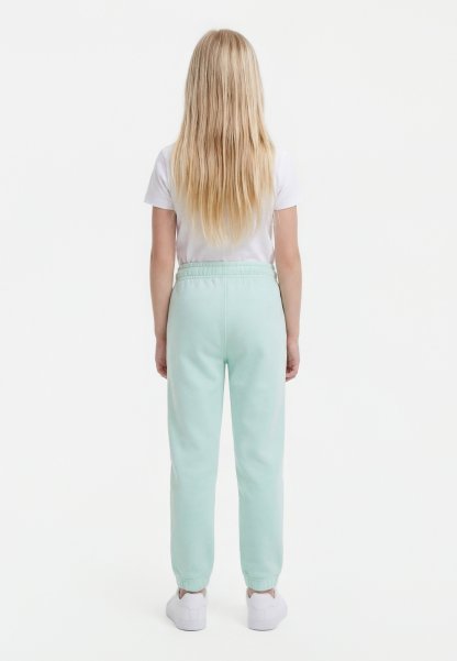 Niño con cabello largo y rubio, vistiendo una camiseta blanca, pantalones de chándal azul claro y zapatillas blancas, de pie de espaldas sobre un fondo blanco.
