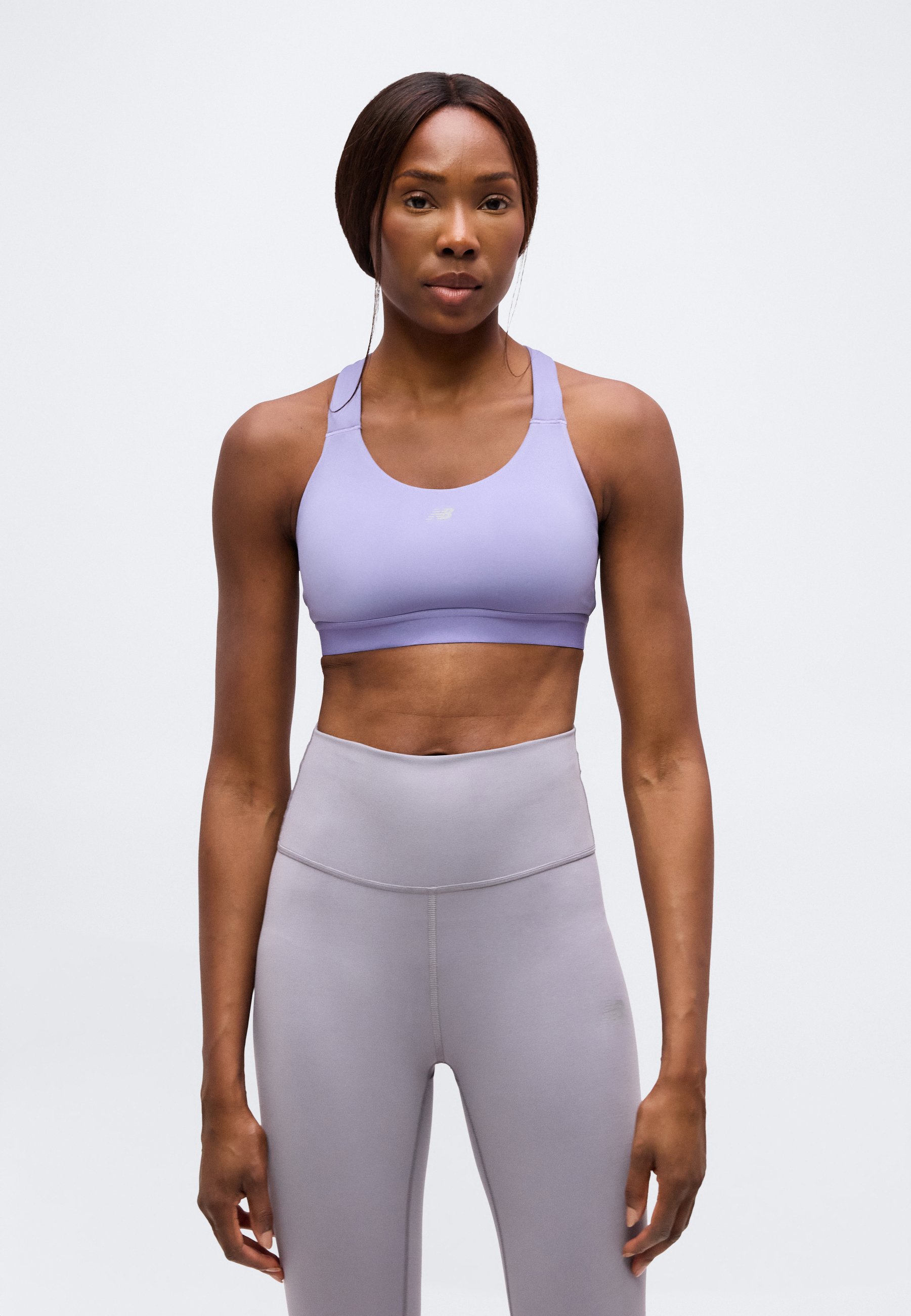 Mujer con piel oscura y cabello recogido, que lleva un sujetador deportivo lavanda y leggings grises de talle alto, de pie frente a un fondo blanco liso.