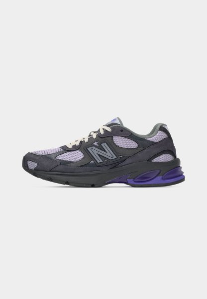 Zapatilla deportiva en tonos gris oscuro y púrpura con tejido de malla, cordones blancos y logotipo "N" de la marca en el lateral, vista de perfil.