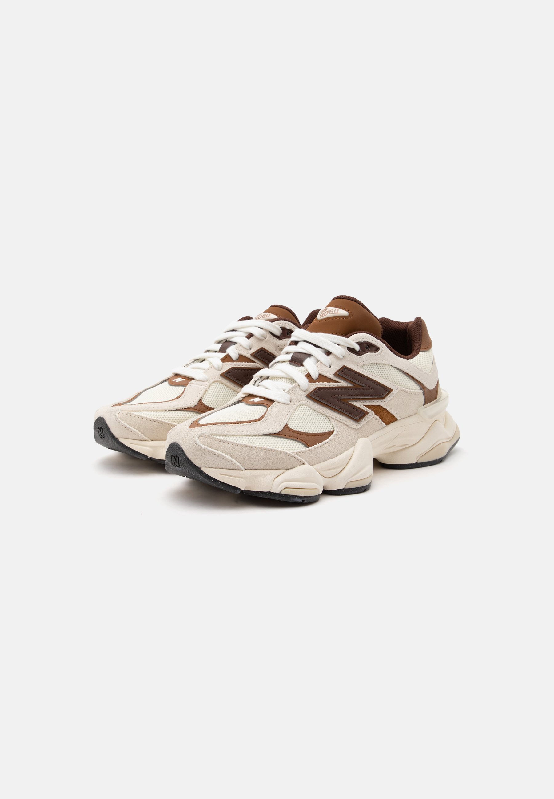 Par de zapatillas New Balance beige y marrón con cordones blancos, detalles de malla y ante, y suelas gruesas y texturizadas sobre un fondo blanco.