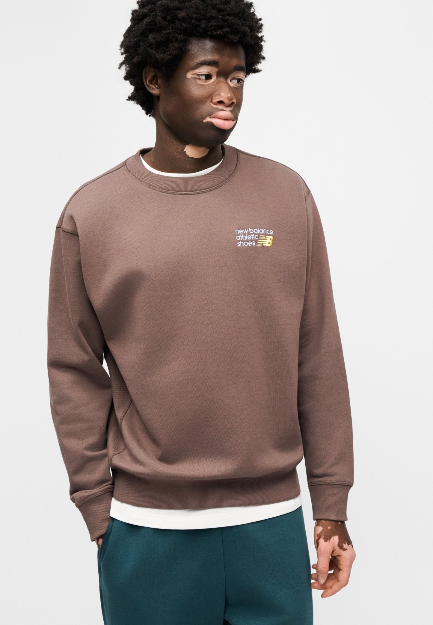 Sudadera marrón con cuello redondo y puños acanalados, con un pequeño logo bordado. Llevada sobre una camisa blanca en capas y combinada con pantalones azul verdoso.