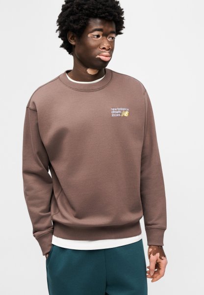 Sudadera marrón con cuello redondo y puños acanalados, con un pequeño logo bordado. Llevada sobre una camisa blanca en capas y combinada con pantalones azul verdoso.
