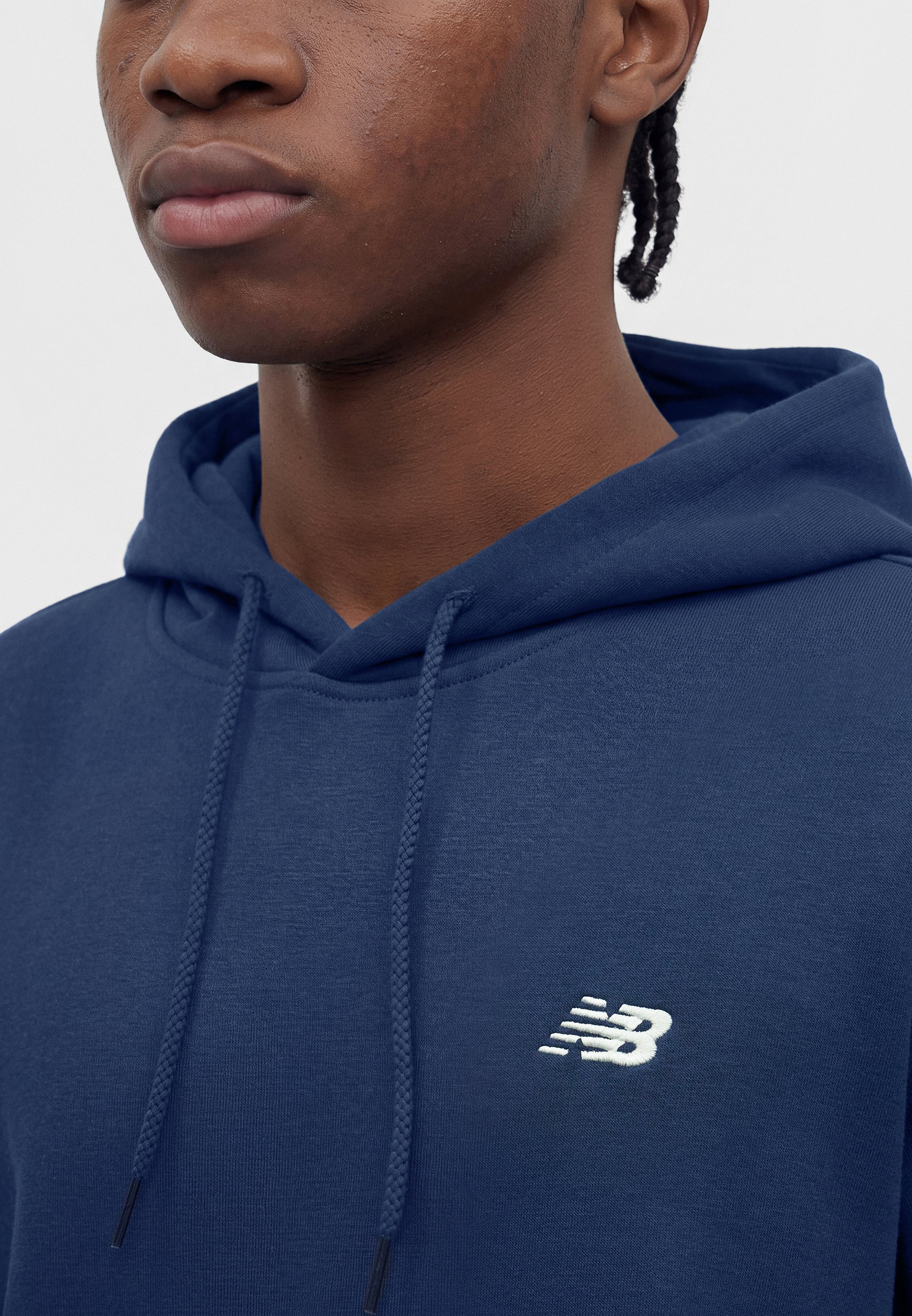 Joven con el pelo trenzado que lleva una sudadera con capucha azul marino de New Balance, mostrando la parte superior del pecho y parte del rostro sobre un fondo liso.
