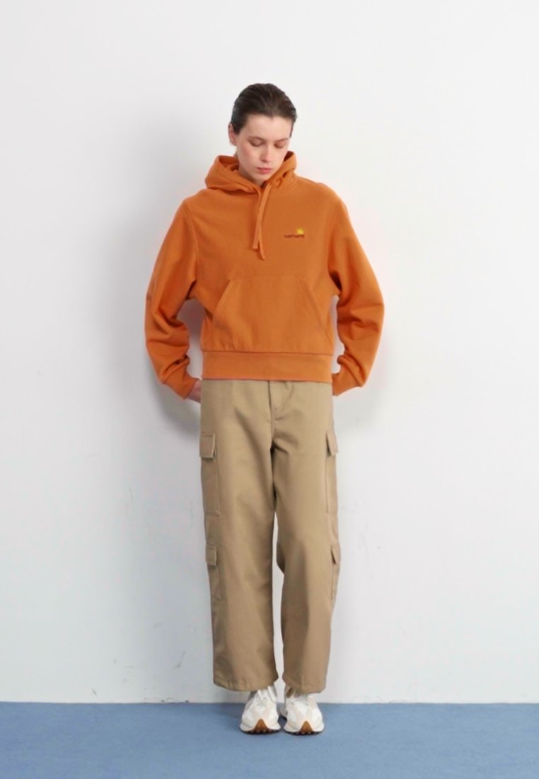 Sudadera naranja con bolsillo delantero y logo, combinada con pantalones cargo beige que tienen bolsillos laterales, y zapatillas blancas. El fondo es liso.