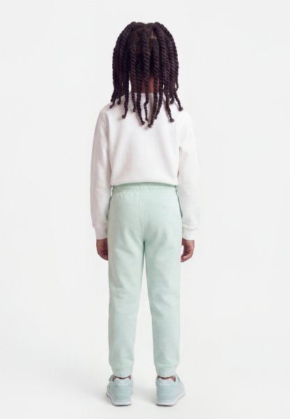 Niño con cabello oscuro trenzado de espaldas, vistiendo una camisa blanca de manga larga, pantalones verdes claros y zapatillas azules claras.