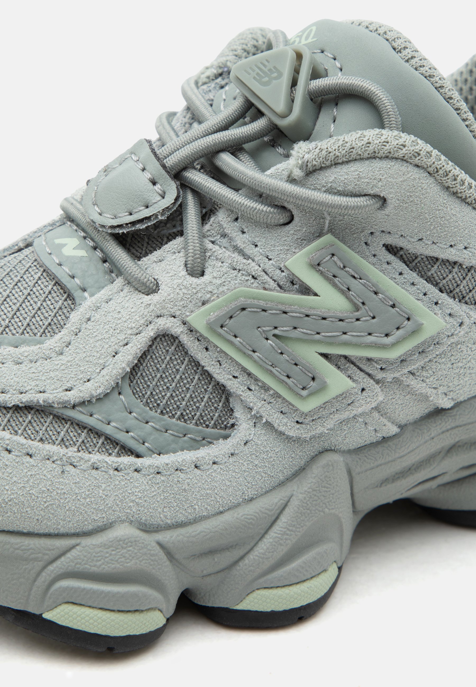 Zapatilla New Balance de color gris claro con paneles de ante y malla, costuras detalladas, cierre con cordones y suela gruesa y texturizada.