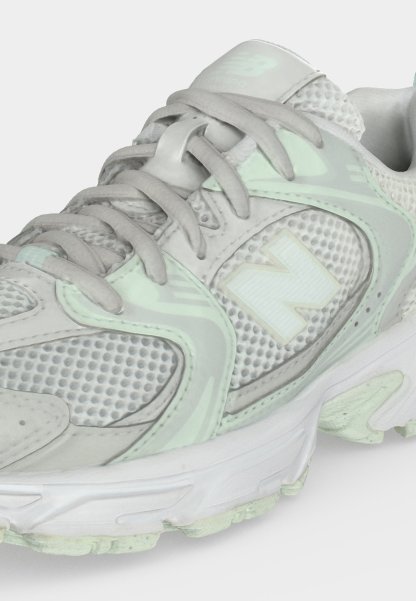 Zapatilla New Balance de color gris claro y verde menta con paneles de malla, cordones y una suela blanca gruesa vista desde un ángulo frontal lateral.