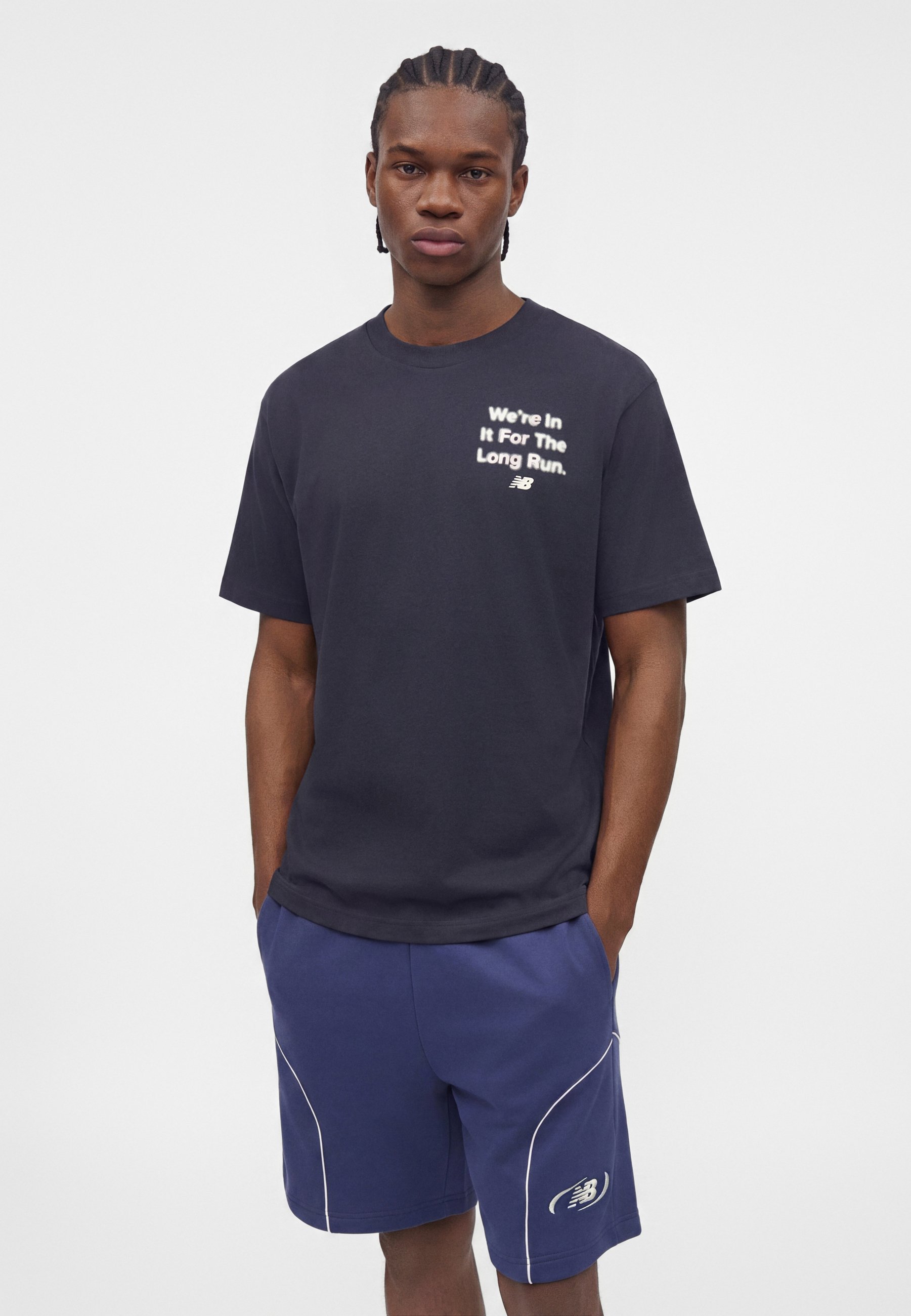 Joven con el pelo trenzado que lleva una camiseta negra con texto blanco y pantalones cortos azules New Balance con ribete blanco, de pie con las manos en los bolsillos.