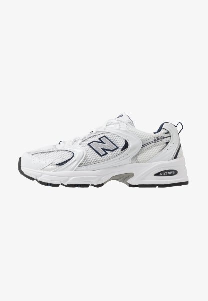 Zapatilla deportiva blanca con parte superior de malla, acentos en azul marino y suela acolchada. Presenta el logo "N", cordones y detalle texturizado en el lateral.
