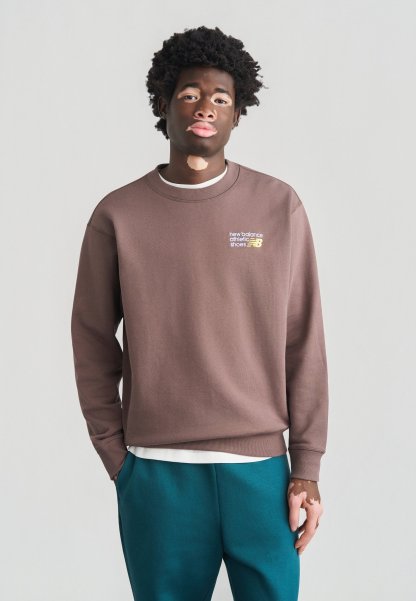 Sudadera marrón con cuello acanalado, con el logo "new balance athletic shoes" en amarillo. Combinada con pantalones azul verdoso. Textura de tela suave.