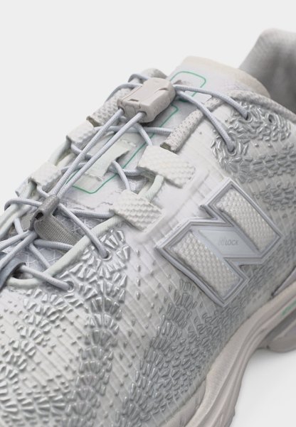 Zapato deportivo blanco con parte superior texturizada, cordones elásticos sin atar asegurados por un taco de plástico y un gran logo "N" en el lateral.
