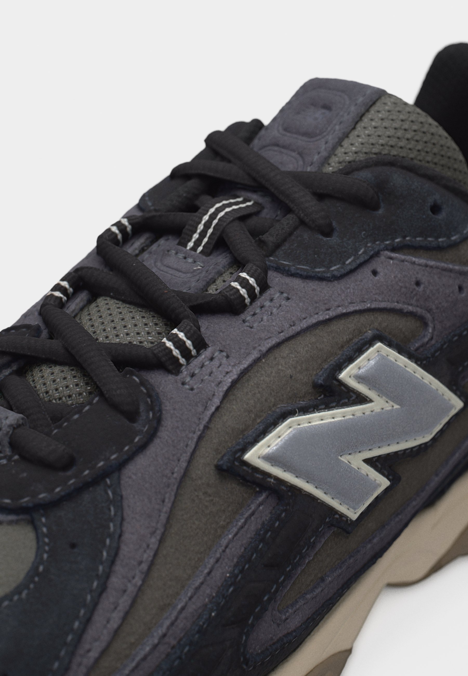 Zapatilla negra y gris con paneles de malla y ante, cordones negros con rayas blancas y un logotipo "N" reflectante en gris en el costado.