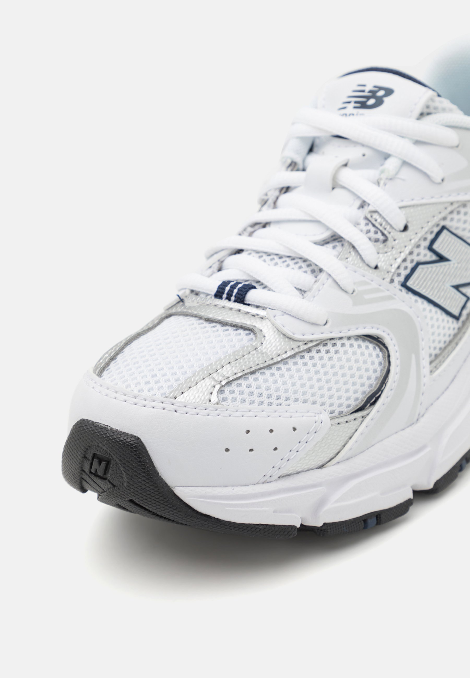 Zapatilla deportiva blanca con parte superior de malla, detalles plateados y suela de goma negra. Incluye un sistema de cordones y un logo visible en el lateral.