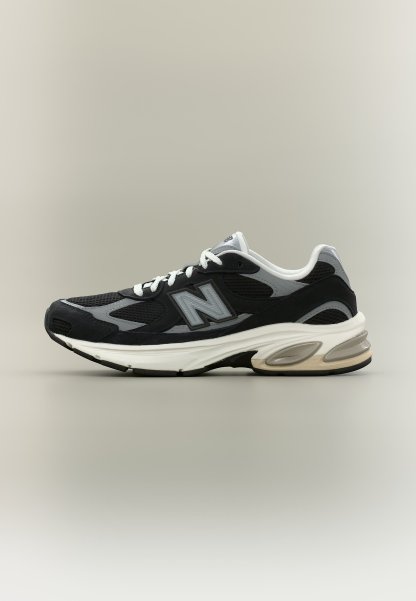 Zapatilla de running New Balance en negro y gris, con cordones blancos y suela gruesa blanca, mostrada de perfil sobre un fondo neutro.