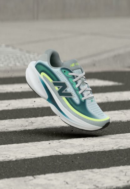 Zapatilla de correr New Balance en gris, azul verdoso y amarillo neón, suspendida sobre un paso de peatones en una carretera asfaltada.