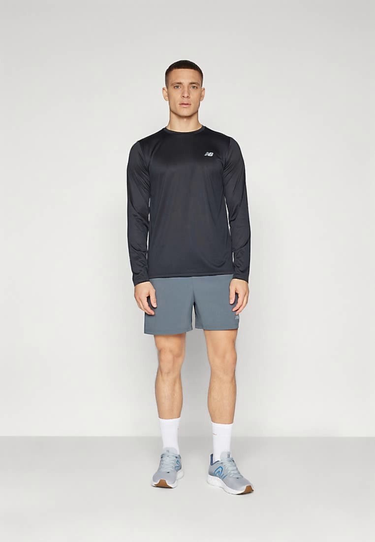 Hombre de pie vistiendo una camiseta atlética de manga larga negra, pantalones cortos grises, calcetas blancas y zapatillas de correr grises contra un fondo liso.