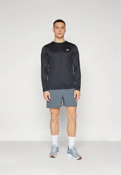 Hombre de pie vistiendo una camiseta atlética de manga larga negra, pantalones cortos grises, calcetas blancas y zapatillas de correr grises contra un fondo liso.