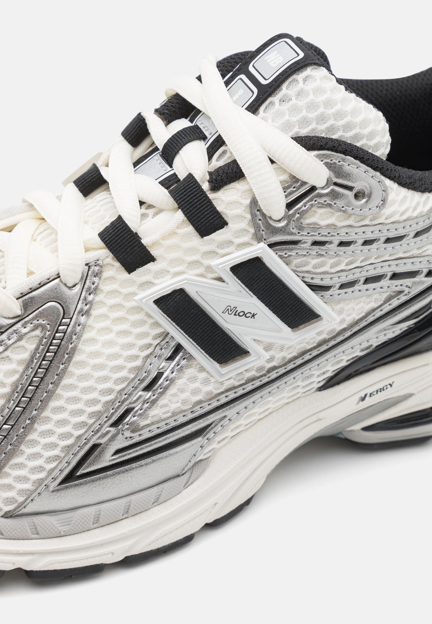 Zapatilla deportiva blanca con parte superior de malla, acentos plateados, cordones negros y una suela texturizada. Presenta el logo 'N' y un diseño acolchado para mayor soporte.