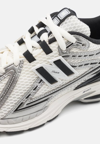 Zapatilla deportiva blanca con parte superior de malla, acentos plateados, cordones negros y una suela texturizada. Presenta el logo 'N' y un diseño acolchado para mayor soporte.