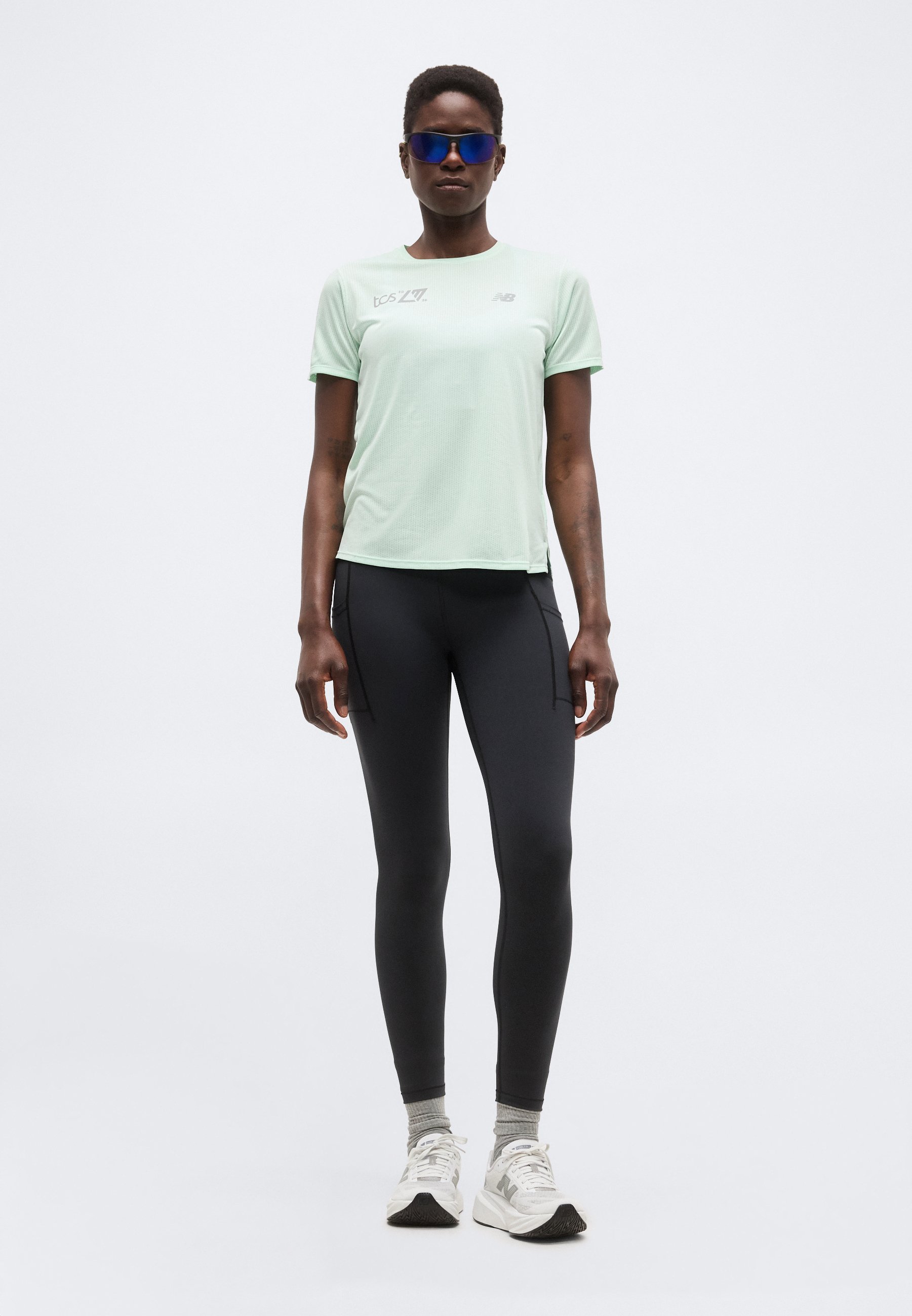Mujer con camiseta de manga corta verde claro, leggings negros, calcetines grises, zapatillas deportivas blancas y gafas de sol deportivas azules de pie frente a un fondo liso.