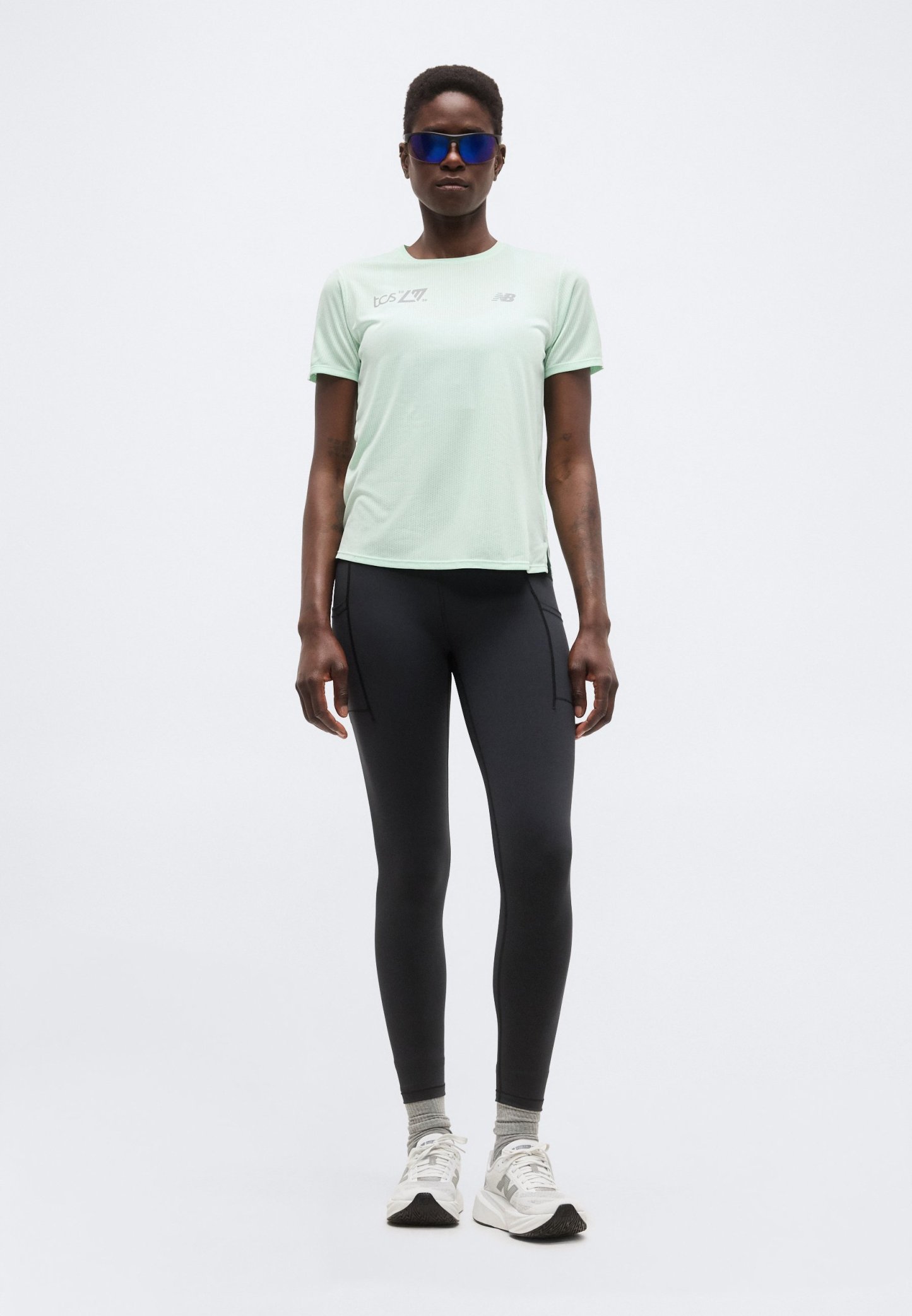 Mujer con camiseta de manga corta verde claro, leggings negros, calcetines grises, zapatillas deportivas blancas y gafas de sol deportivas azules de pie frente a un fondo liso.
