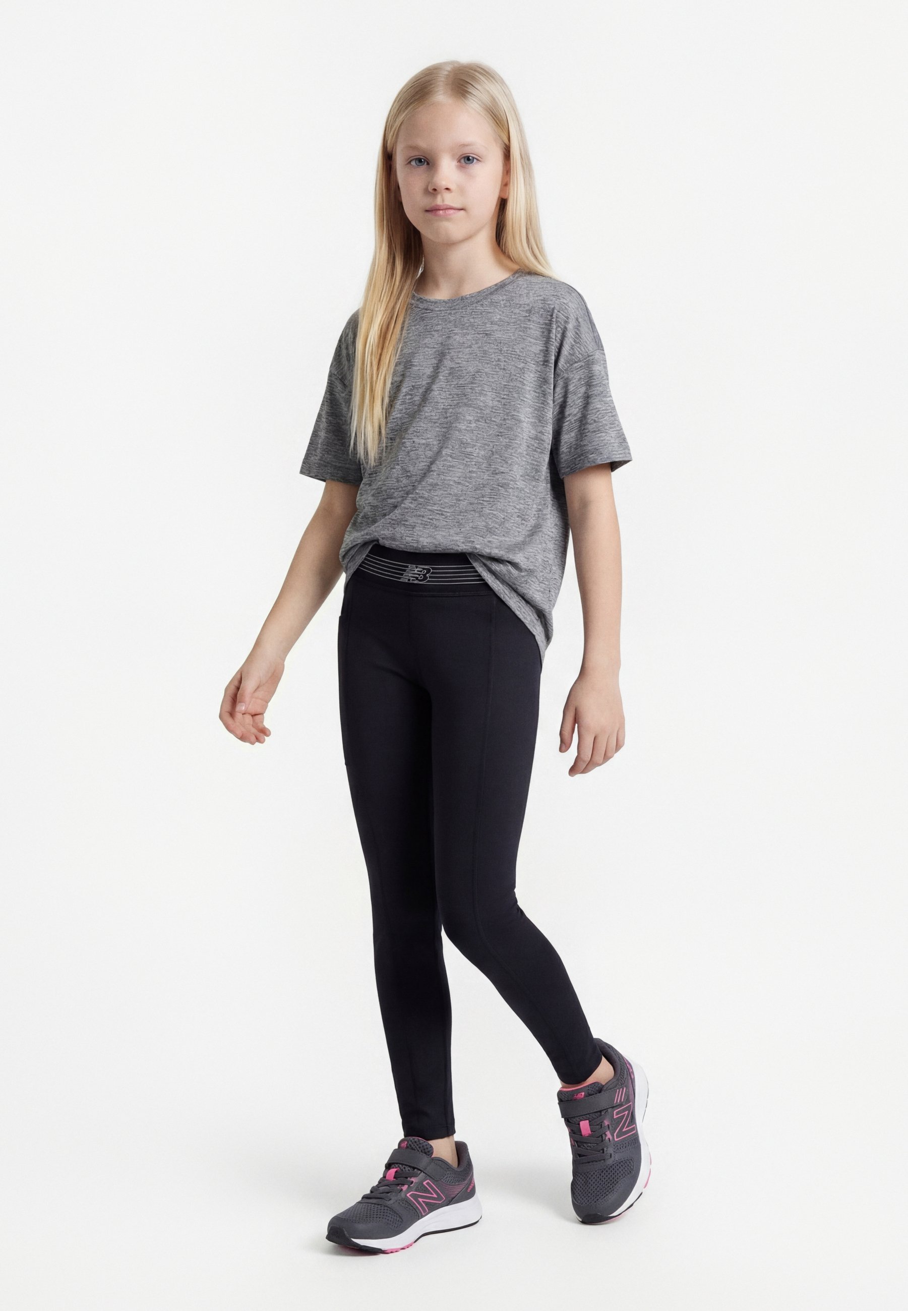 Niña joven de pie, vestida con una camiseta gris, leggings negros y zapatillas deportivas grises y rosas sobre un fondo blanco.