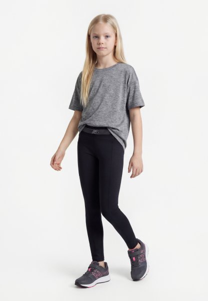Niña joven de pie, vestida con una camiseta gris, leggings negros y zapatillas deportivas grises y rosas sobre un fondo blanco.