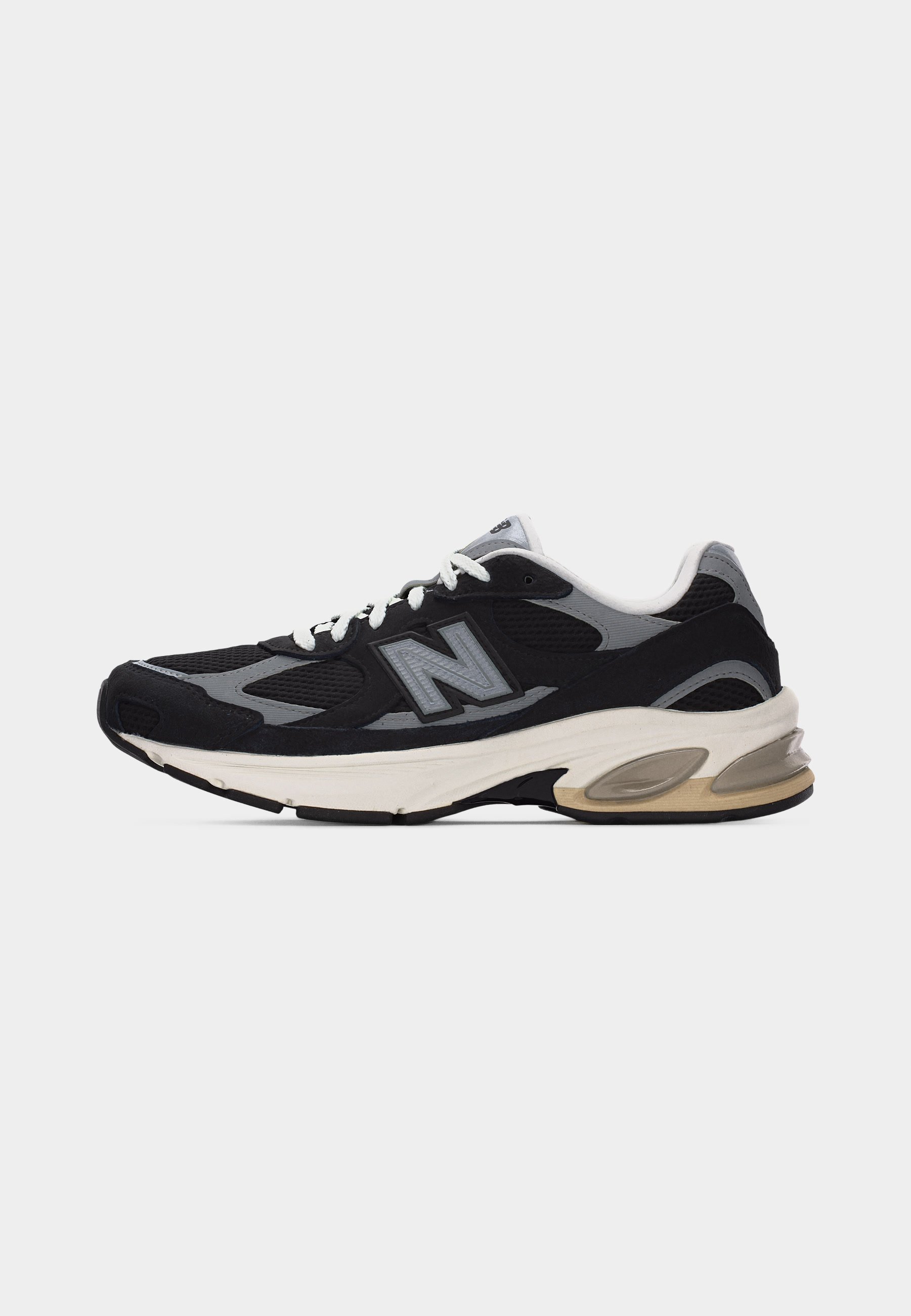 Zapatilla deportiva negra y gris con suela blanca, cordones blancos y un gran logotipo "N" en el costado, vista de perfil lateral.