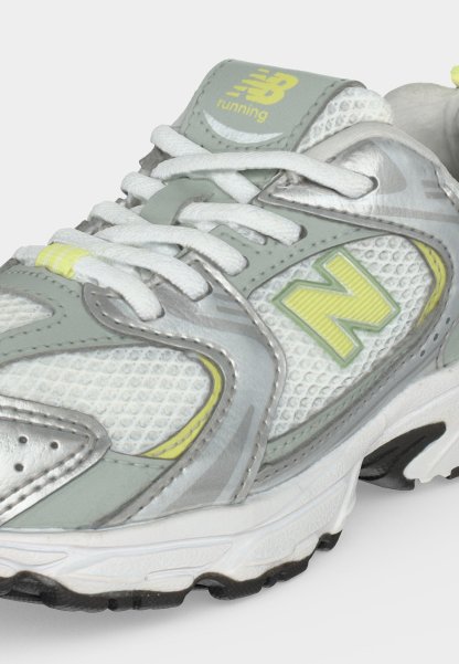Zapatilla de running New Balance en gris, blanco y amarillo con tejido de malla, cordones y suela texturizada, mostrada de cerca.