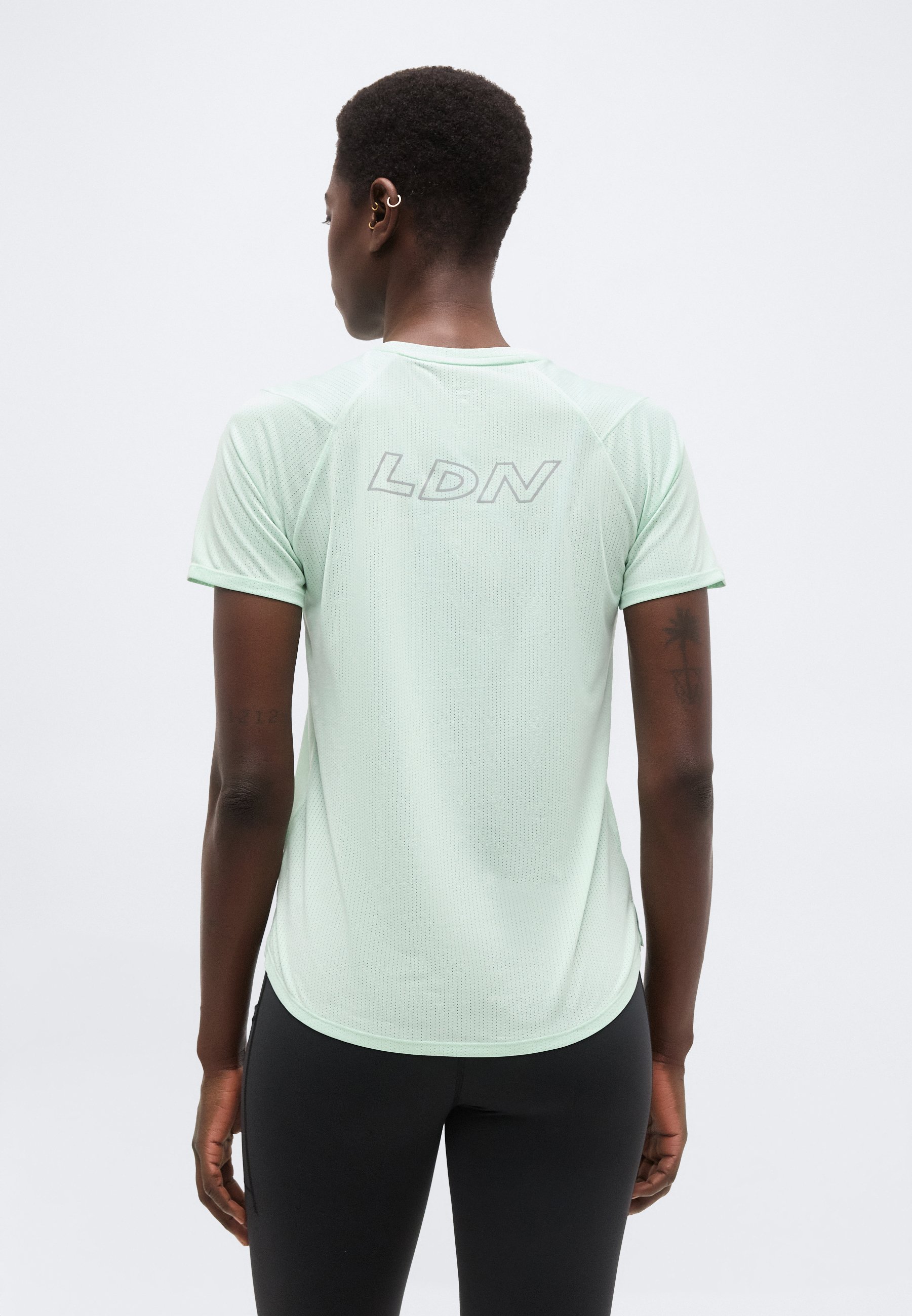 Persona con cabello corto que lleva una camiseta de malla de manga corta verde claro con "LDN" en la espalda y leggings negros, de espaldas.