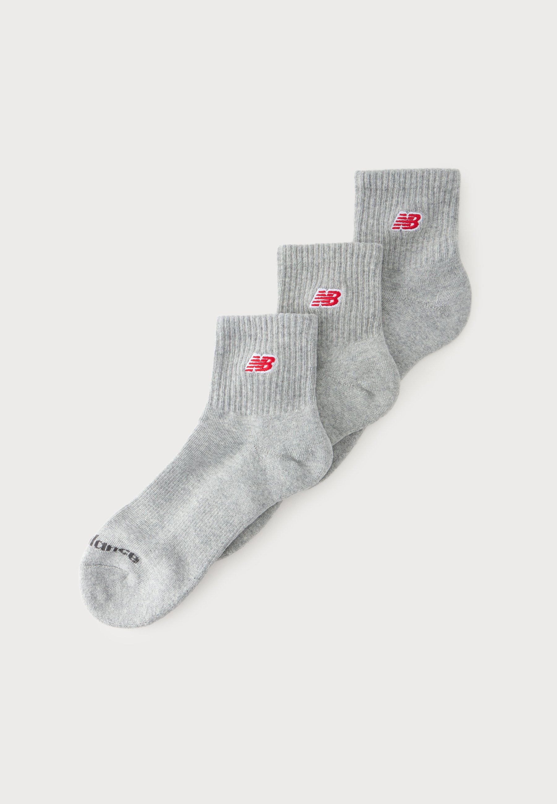 Tres pares de calcetines tobilleros grises con puños acanalados y logotipos rojos de New Balance están apilados sobre un fondo blanco.